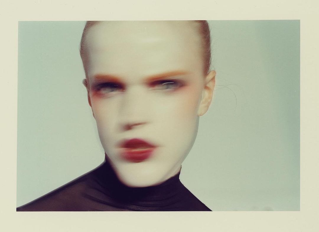 Portrait of androgynous person with striking makeup and shaved head, wearing a black turtleneck shirt against a light background.