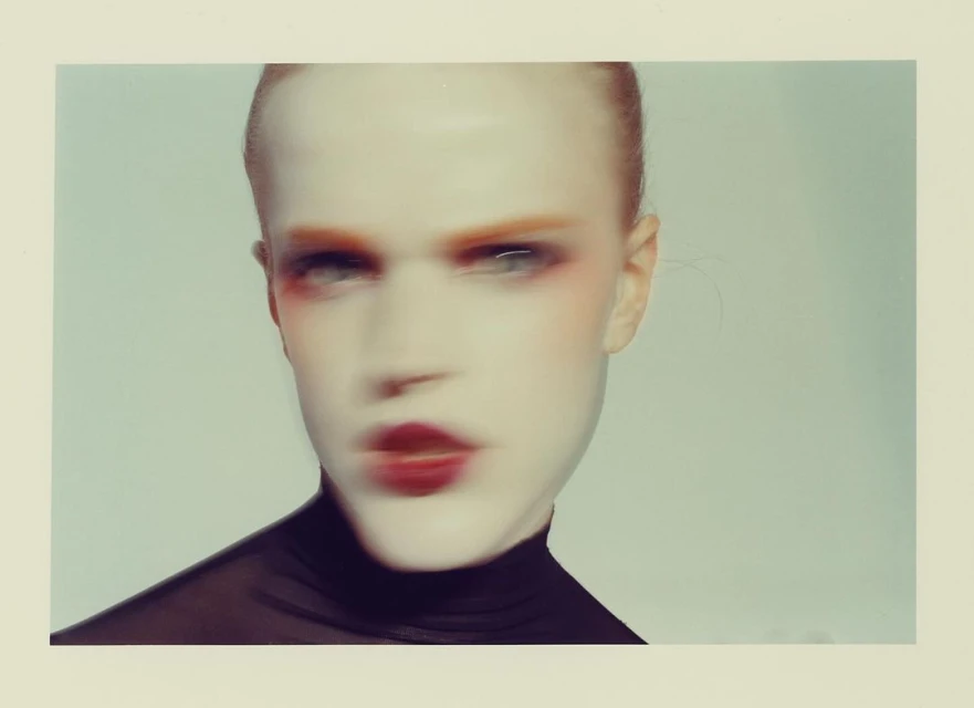 Portrait of androgynous person with striking makeup and shaved head, wearing a black turtleneck shirt against a light background.