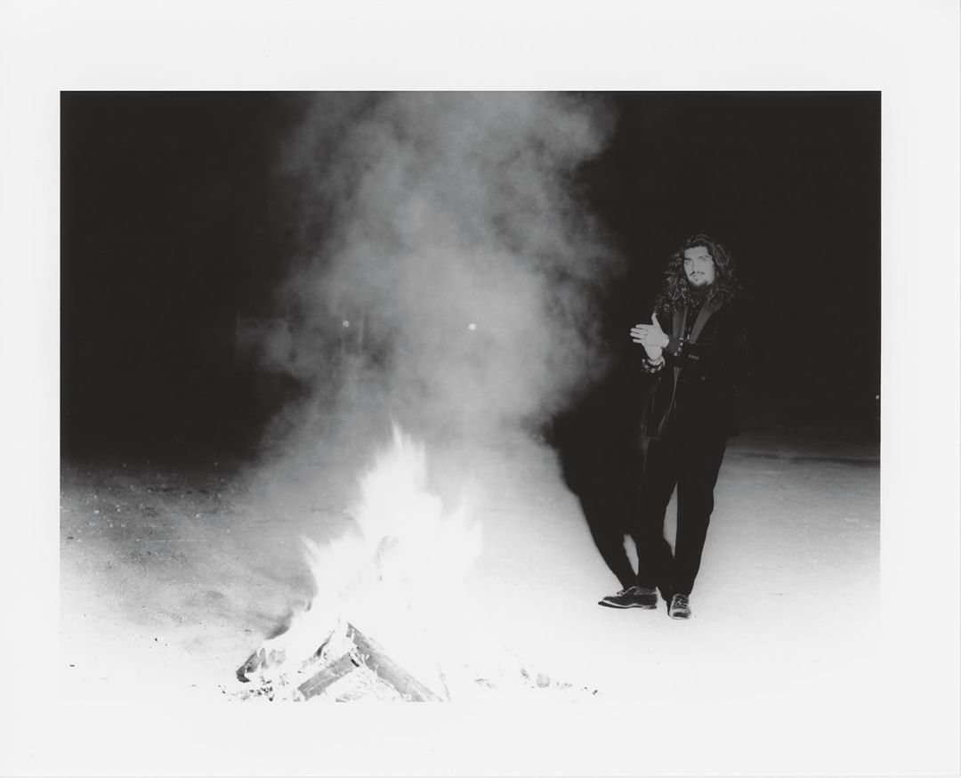 Adult male standing outside next to a burning pile of wood creating a large plume of smoke on a dark night with a spotlight glow.