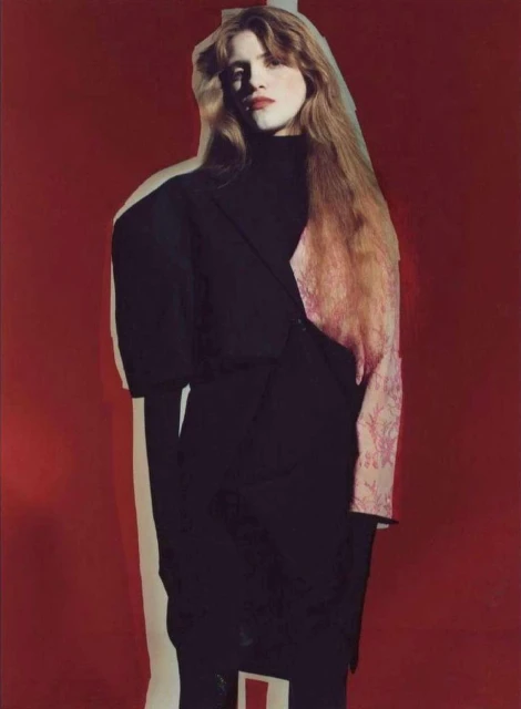 Fashion editorial shot of a woman with long red hair wearing a black dress against a bold red background with a cutout shadow.