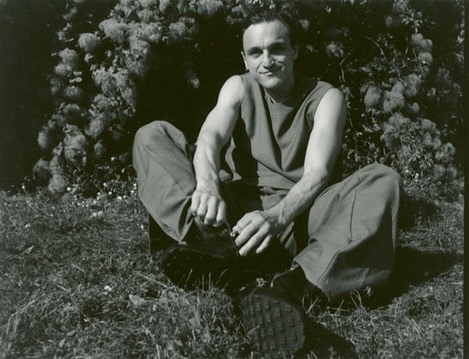Man in tank top and loose pants sits on grass in front of flowering bushes, smoking a cigarette, with black and white aesthetic.