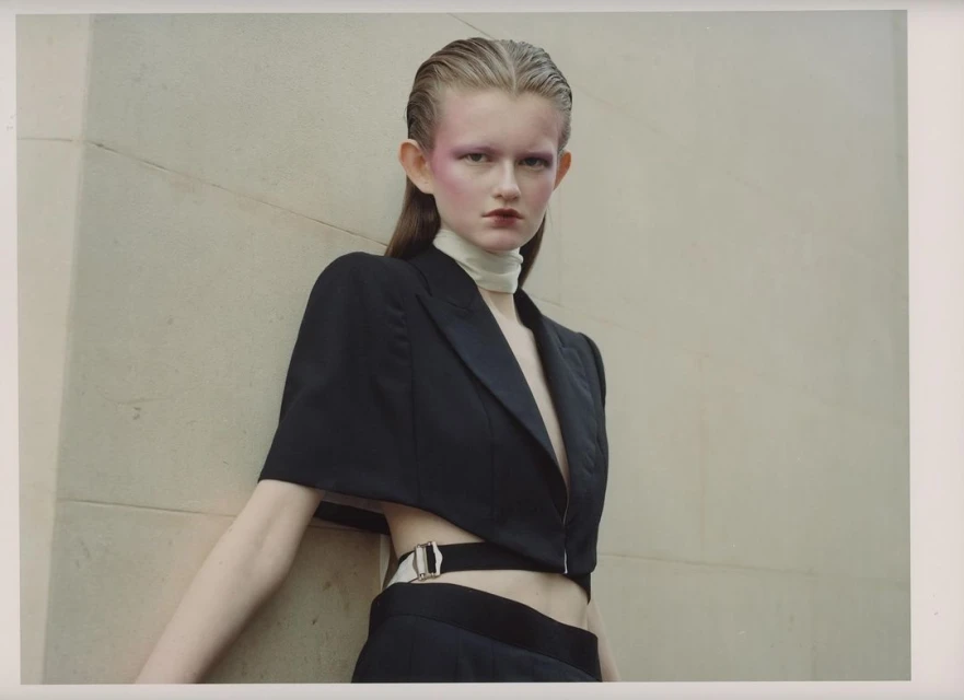 Fashion-forward model poses against a neutral wall in a cropped black jacket, showcasing bold eye makeup and slicked-back hair.