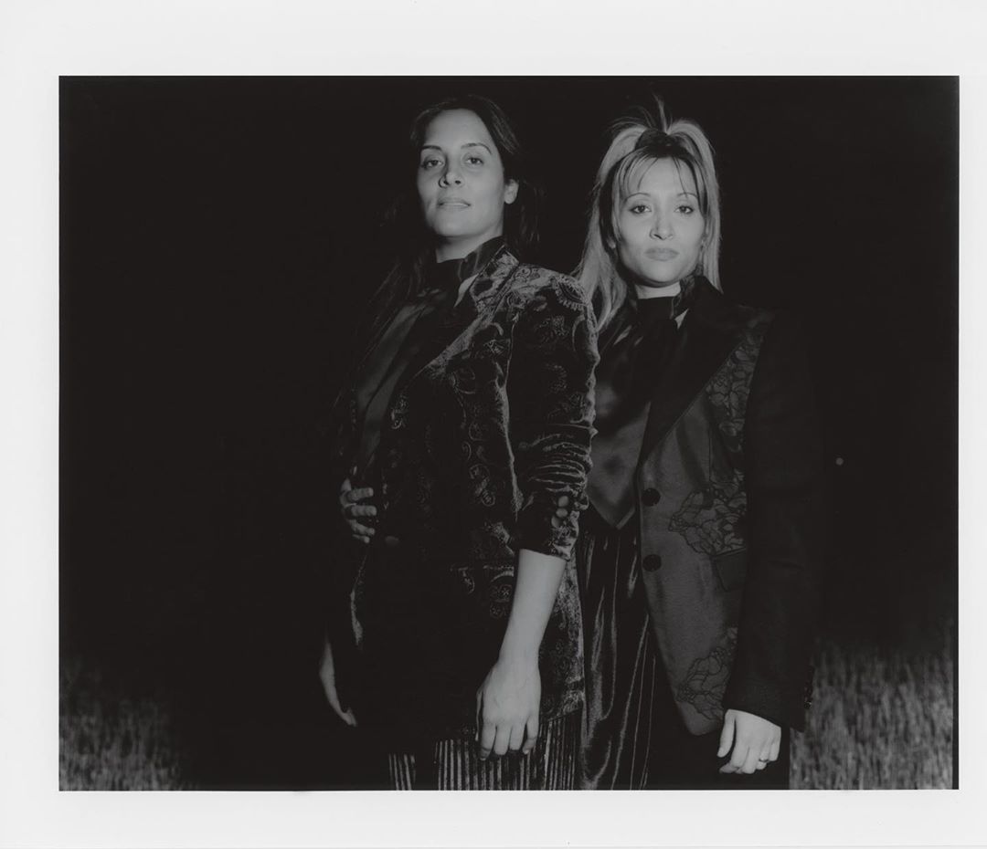 Two adult women pose in velvet jackets against a dark background in a black and white photograph with shallow depth of field.
