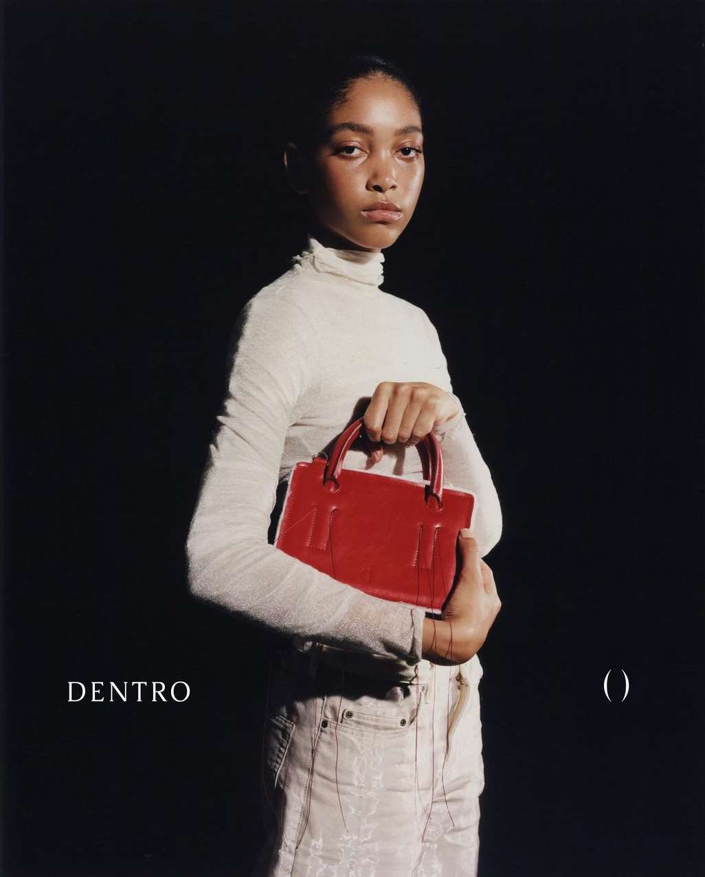 Woman poses with a bright red handbag. She wears a cream-colored turtleneck and light jeans against a solid black backdrop in studio lighting.