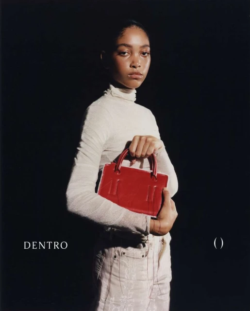 Woman poses with a bright red handbag. She wears a cream-colored turtleneck and light jeans against a solid black backdrop in studio lighting.