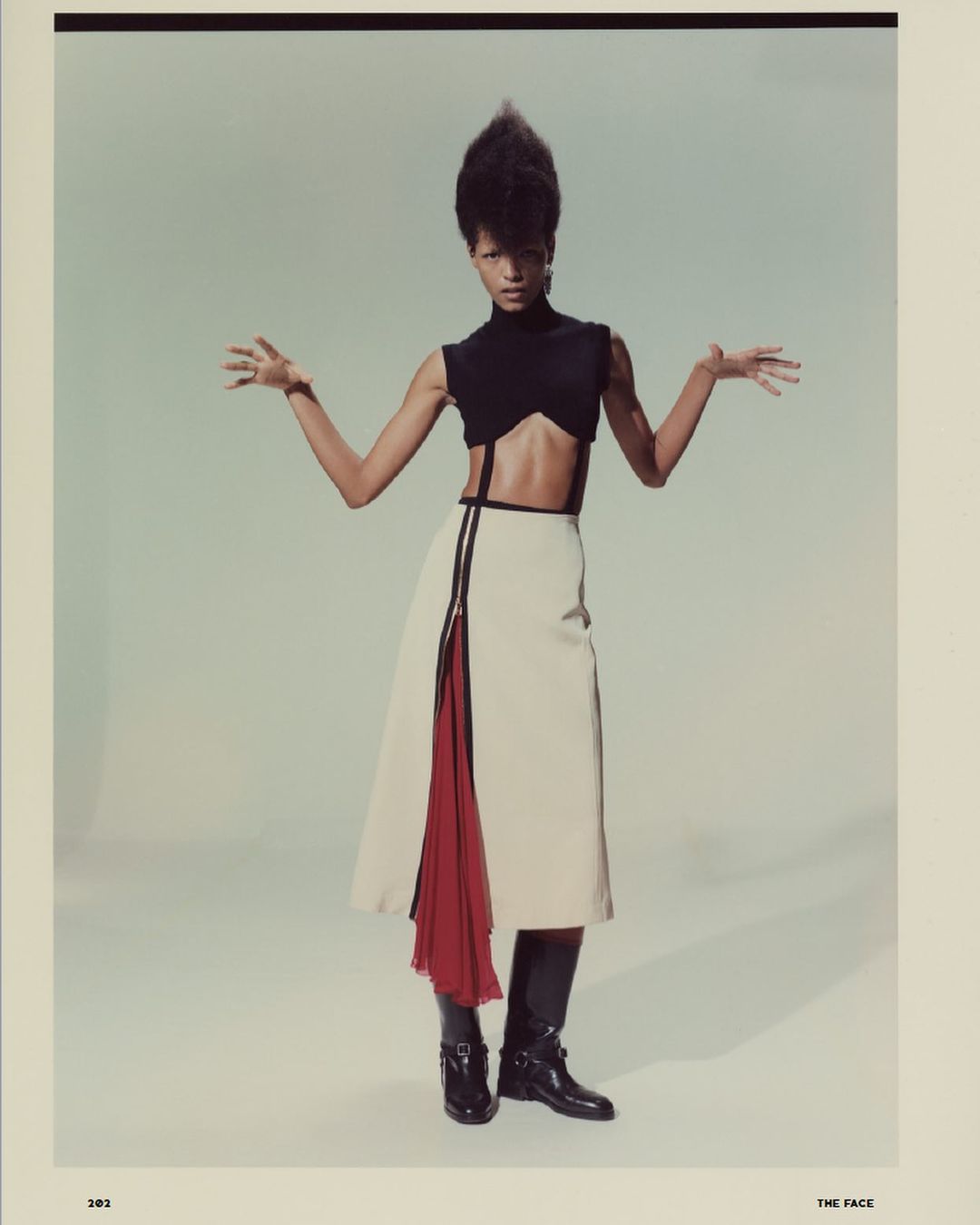 Fashion model poses in black crop top, tan skirt with red accent, and black boots in a studio setting with neutral lighting.