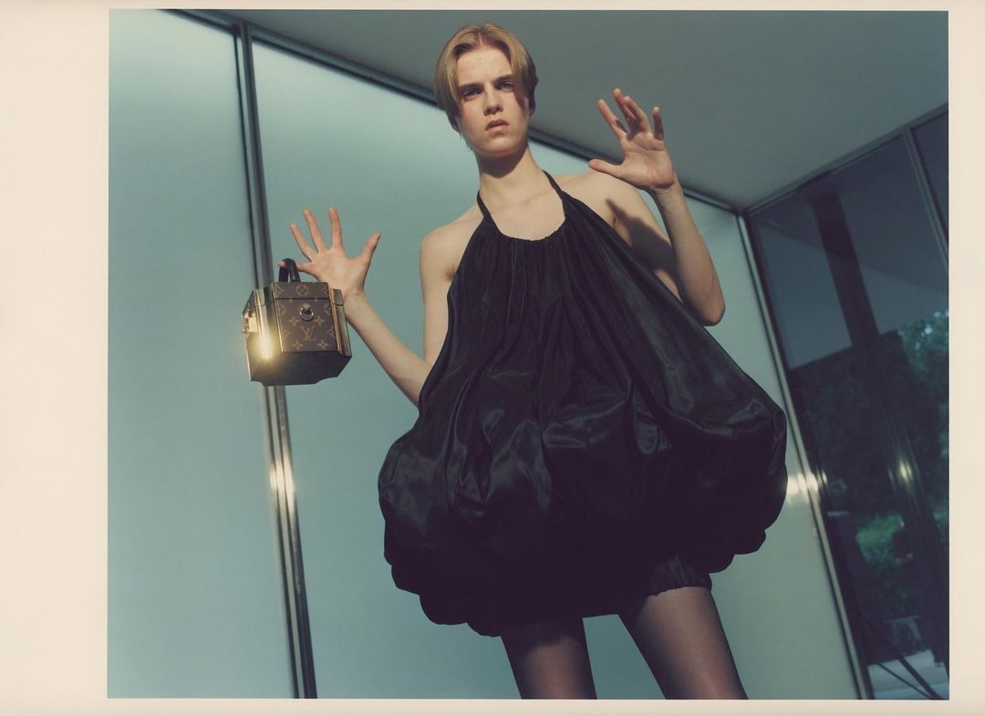 Young woman in a black dress poses with a decorative box in an indoor space with large windows and a concrete floor, showcasing fashion and style.