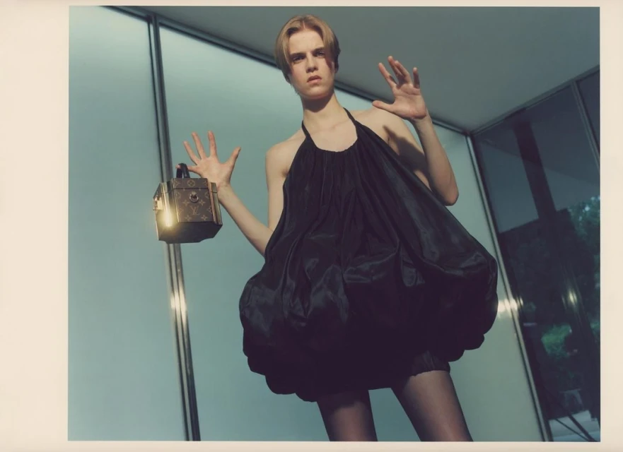 Young woman in a black dress poses with a decorative box in an indoor space with large windows and a concrete floor, showcasing fashion and style.