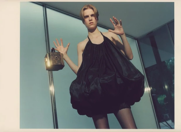 Young woman in a black dress poses with a decorative box in an indoor space with large windows and a concrete floor, showcasing fashion and style.