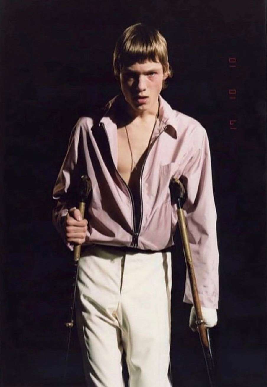 A person with crutches poses in a pink jacket and cream pants against a dark background, illuminated by a spotlight.