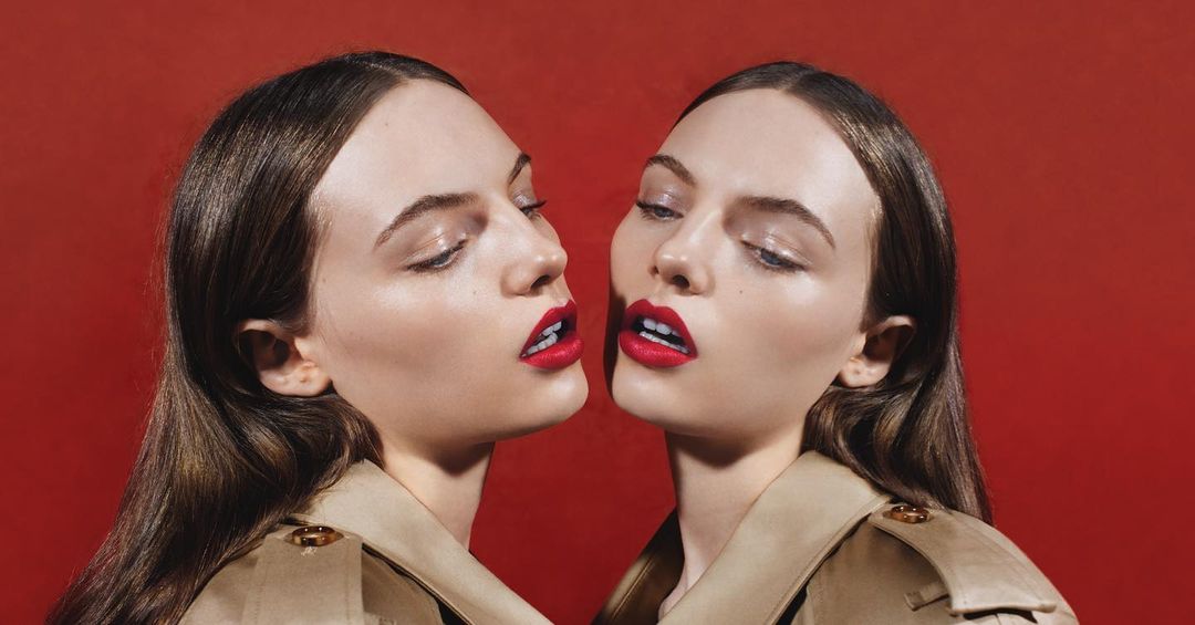 Close-up studio shot of two young adult women with matching brunette hair, tan trench coats, and bright red lipstick against a red backdrop.