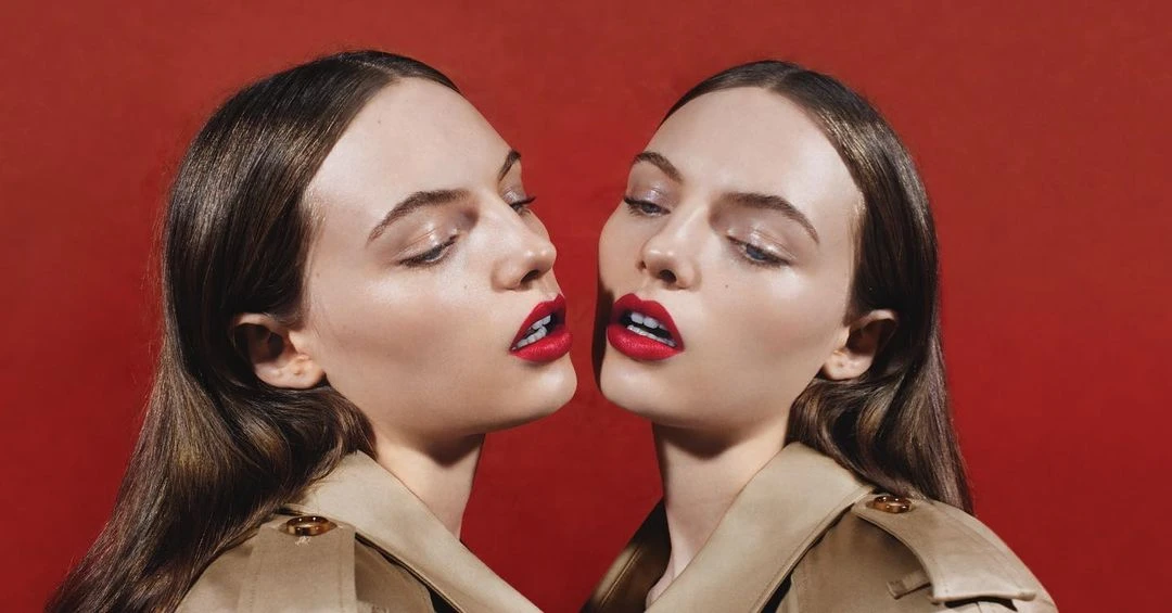 Close-up studio shot of two young adult women with matching brunette hair, tan trench coats, and bright red lipstick against a red backdrop.