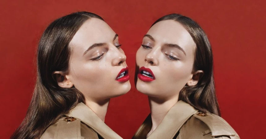 Close-up studio shot of two young adult women with matching brunette hair, tan trench coats, and bright red lipstick against a red backdrop.