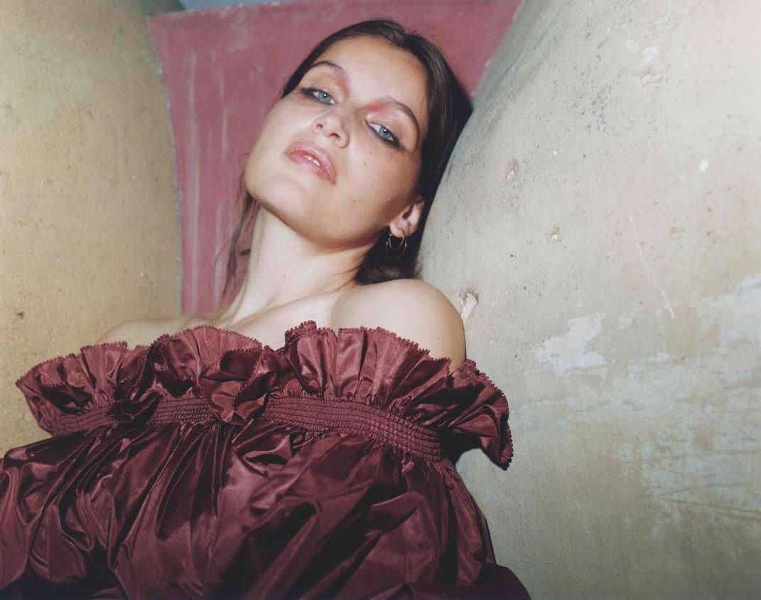 Woman with dark hair and smokey eyes wearing a maroon ruffled dress in a close-up shot against a textured wall in a beauty portrait.