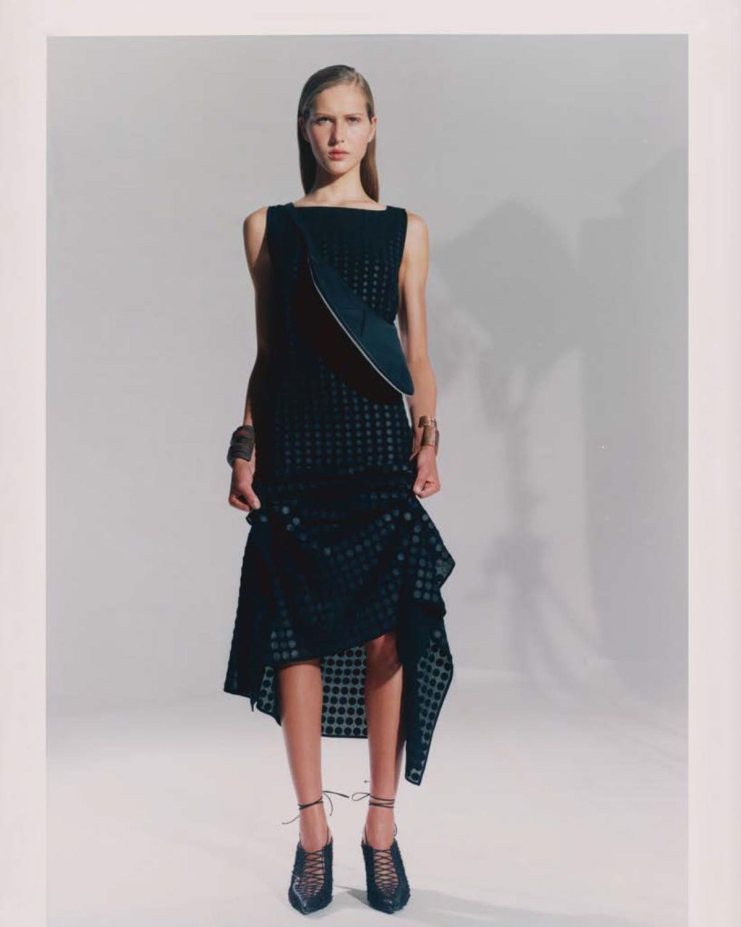Fashion model poses in a black sleeveless dress with asymmetrical hem in a studio setting with soft lighting and neutral background.