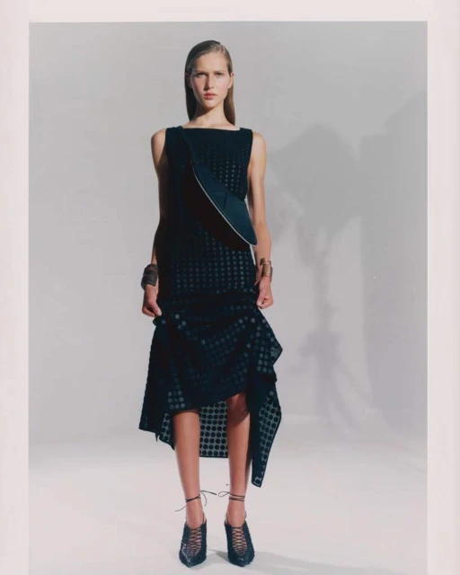 Fashion model poses in a black sleeveless dress with asymmetrical hem in a studio setting with soft lighting and neutral background.