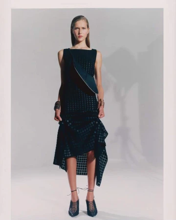 Fashion model poses in a black sleeveless dress with asymmetrical hem in a studio setting with soft lighting and neutral background.
