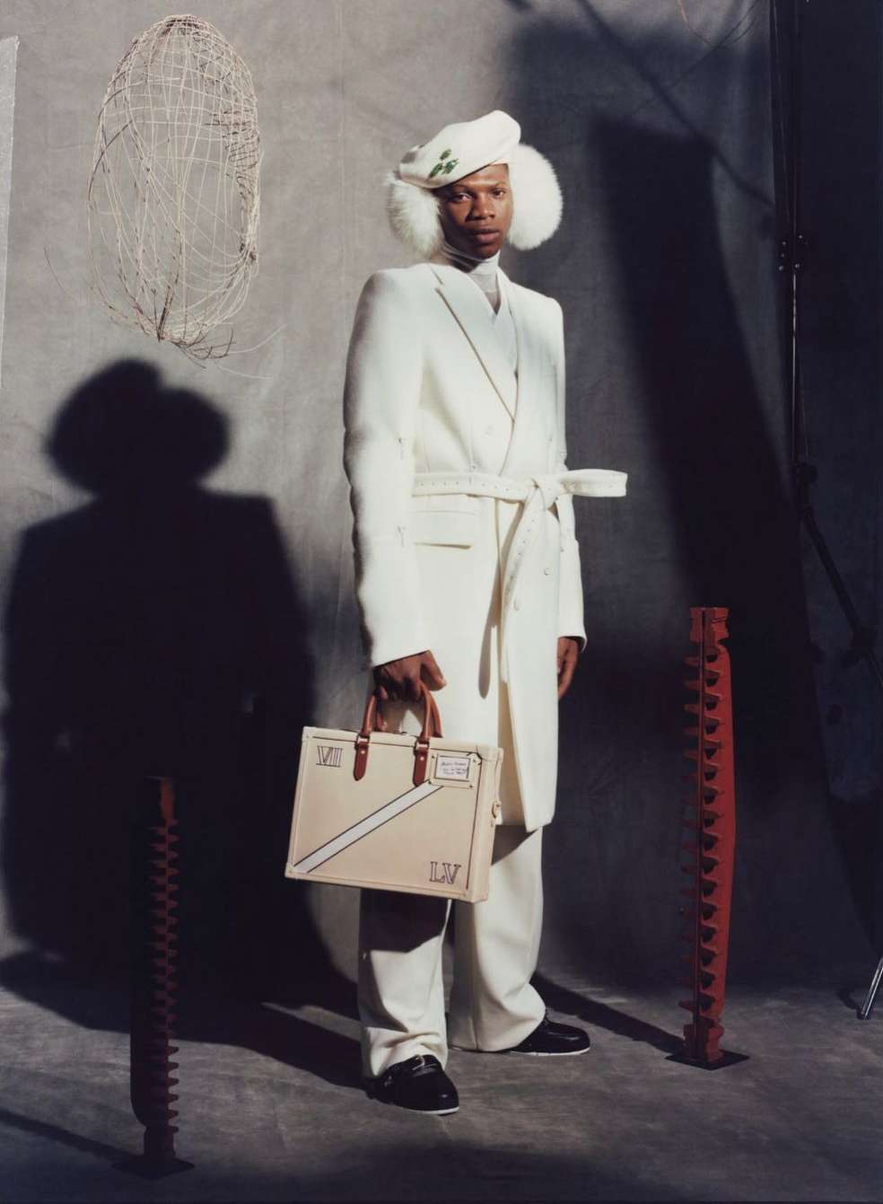 Full shot of an adult male in a white coat, pants, and hat, holding a beige handbag in a studio setting with props.
