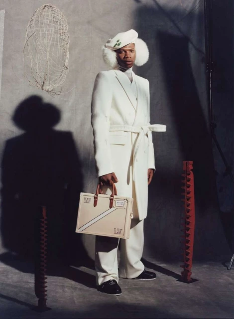 Full shot of an adult male in a white coat, pants, and hat, holding a beige handbag in a studio setting with props.