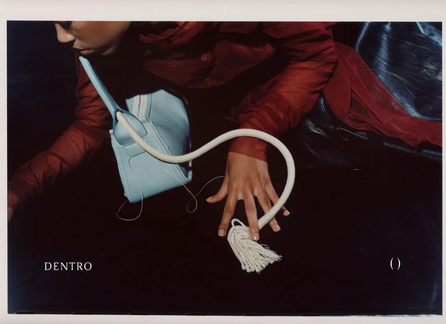 Fashion editorial shot of a light-blue handbag with white handle tassel, being held by a model in a red top against a dark background.