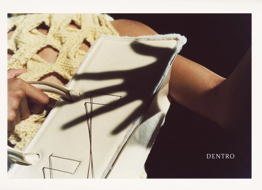 Close-up on a light yellow knit sweater and off-white purse with handle held by a person with a hand shadow over the purse.