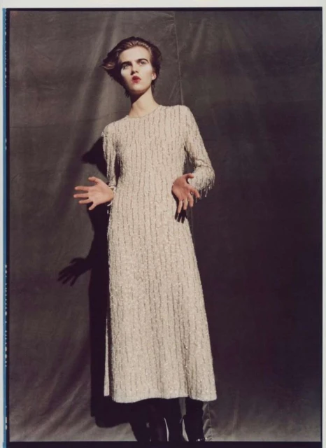 Fashion shot of a young fair-skinned woman in a sparkling gown, with dramatic makeup, against a dark grey backdrop in a photo studio.