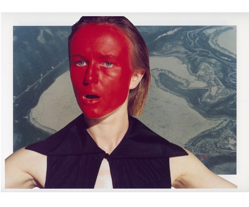 Portrait of a woman with red face paint, wearing a black top, posing in front of a print featuring an aerial landscape.