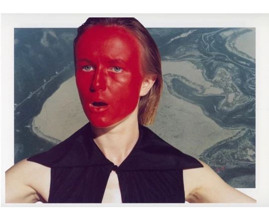 Portrait of a woman with red face paint, wearing a black top, posing in front of a print featuring an aerial landscape.