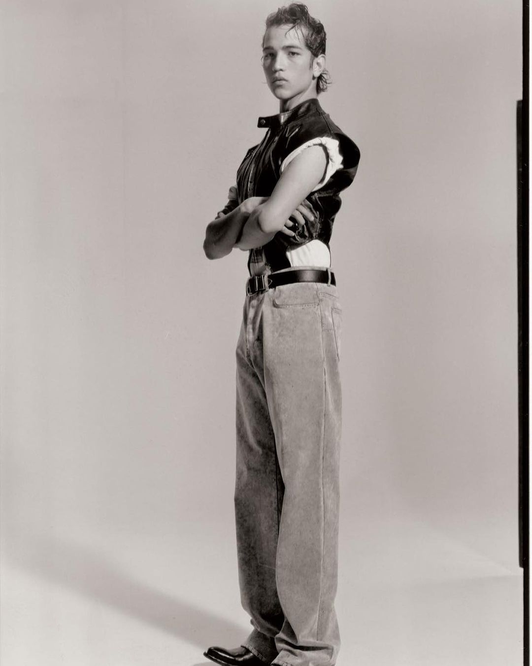 Young man in a vest poses with arms crossed in a studio against a plain background, wearing wide leg pants and black shoes in a full shot.