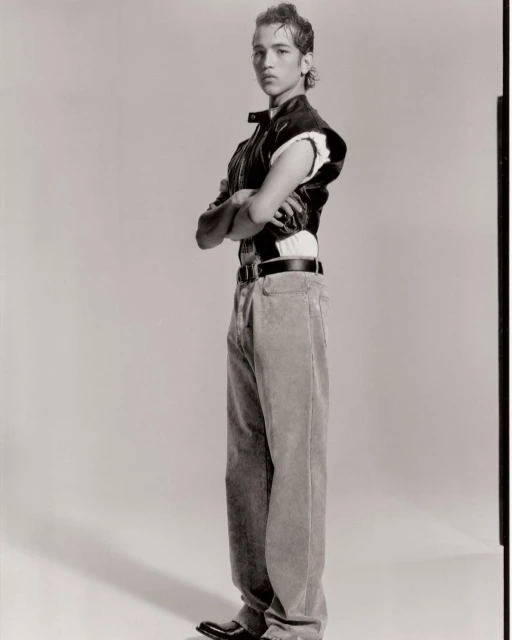 Young man in a vest poses with arms crossed in a studio against a plain background, wearing wide leg pants and black shoes in a full shot.