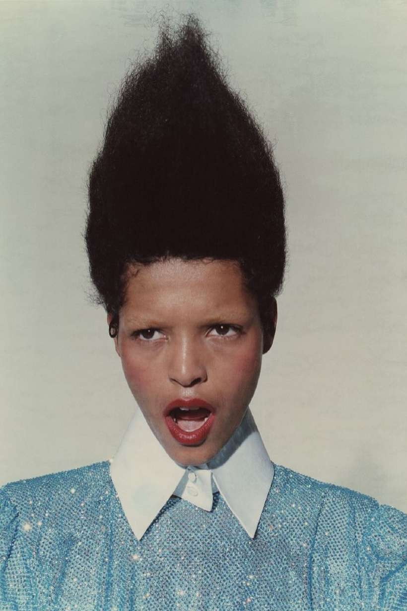 Portrait of woman with high, dark afro hairstyle wearing a sparkling blue dress with white collar against a pale background.