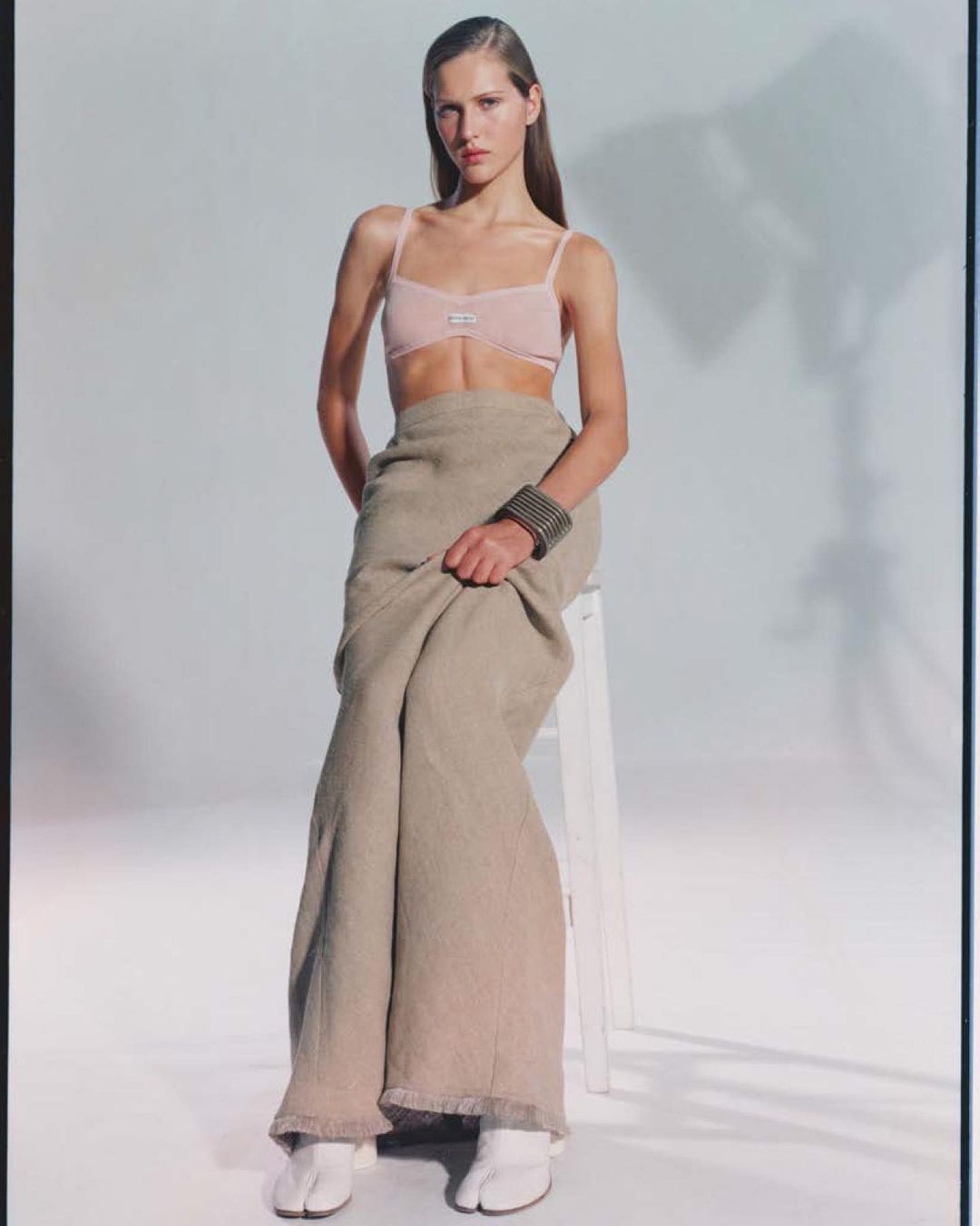 Elegant female fashion model poses in a light pink bra, flowing skirt, and white footwear in a bright photo studio with soft lighting.