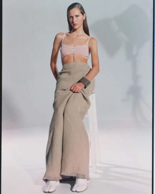 Elegant female fashion model poses in a light pink bra, flowing skirt, and white footwear in a bright photo studio with soft lighting.