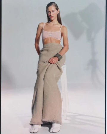 Elegant female fashion model poses in a light pink bra, flowing skirt, and white footwear in a bright photo studio with soft lighting.