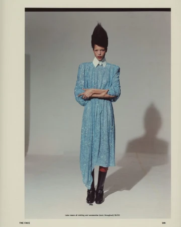 Model with prominent spiky hair wearing a blue textured dress and black boots against a plain grey backdrop
