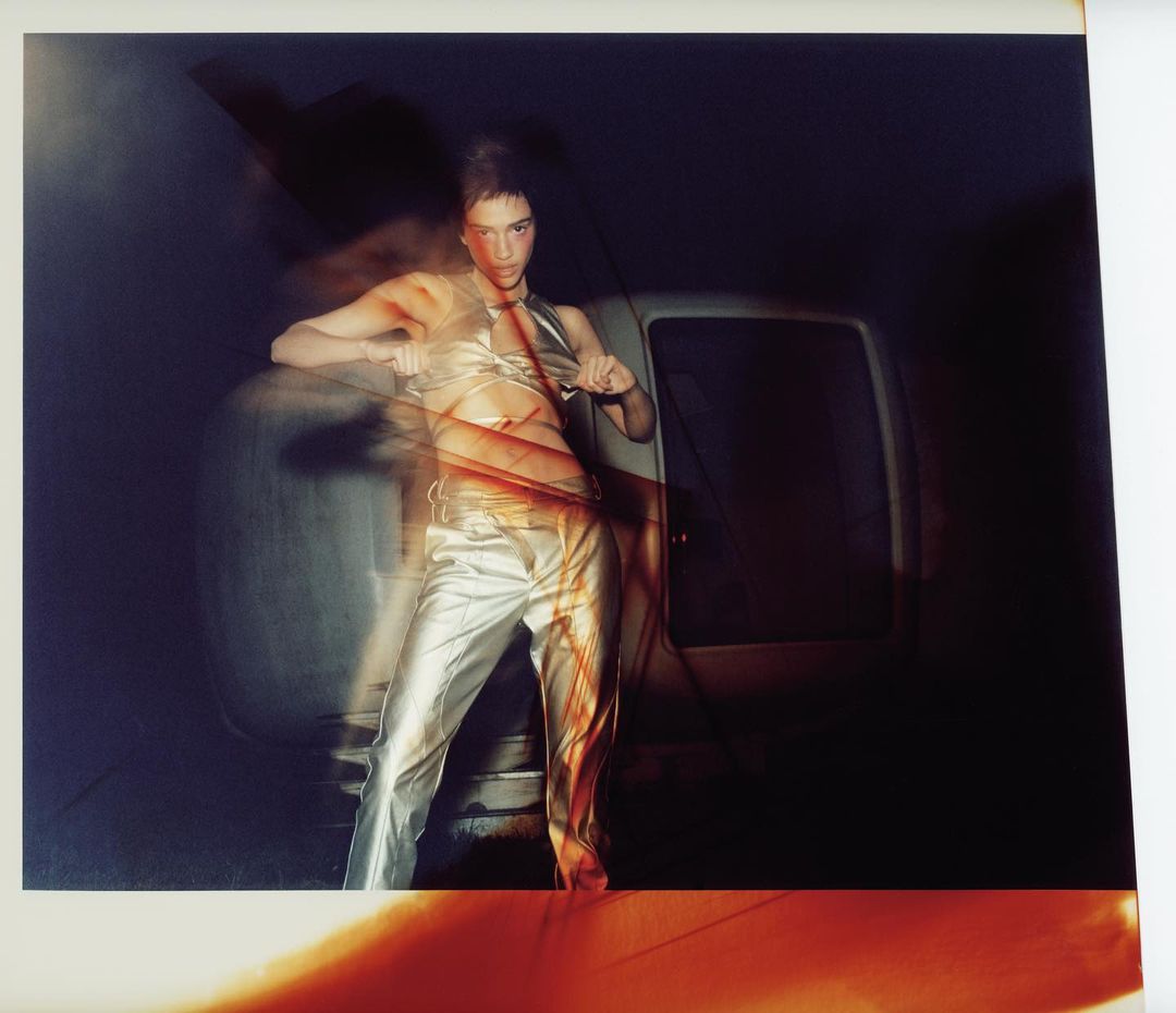 Adult female in a silver outfit leans against a car with a blurred, dreamlike effect. Fashion photography with a dark, edgy aesthetic.