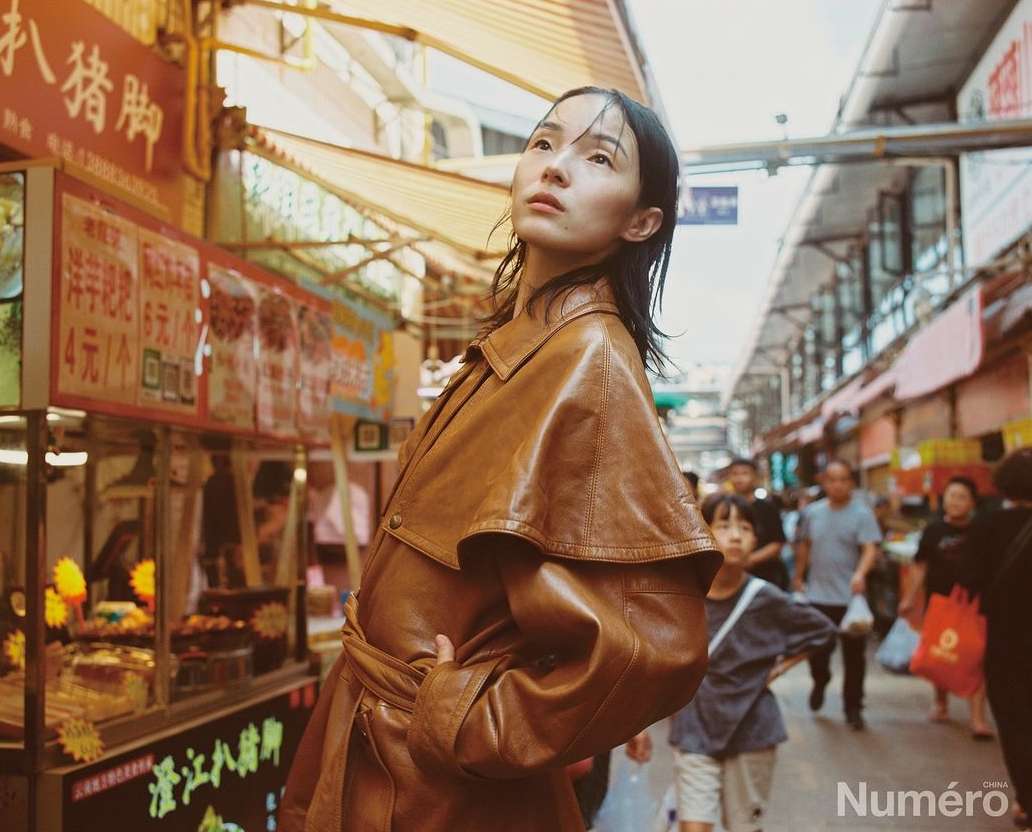 Adult woman in a brown leather coat looking up in an outdoor market with food stands and people in the background, fashion editorial.