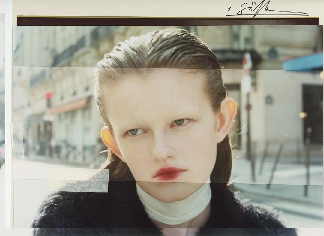 Close-up studio-style portrait of a young woman with slicked-back hair and dark coat against a blurred urban street backdrop.