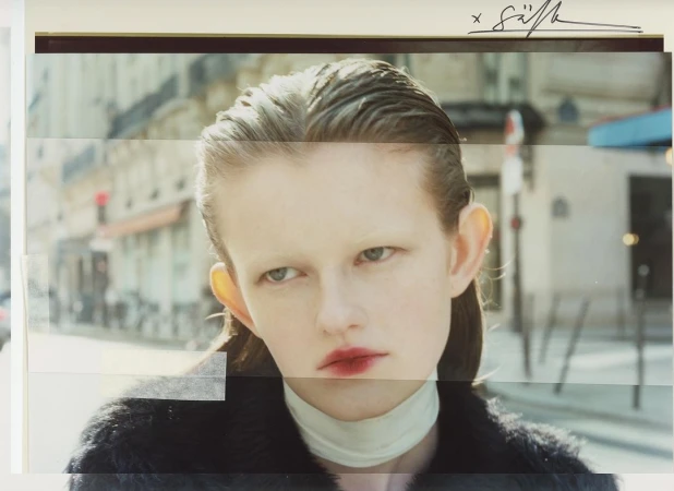 Close-up studio-style portrait of a young woman with slicked-back hair and dark coat against a blurred urban street backdrop.