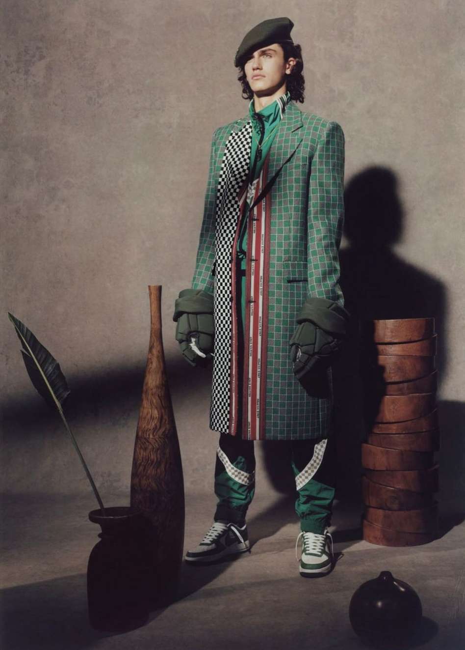 Fashion shot of a male model wearing a green checkered coat, cap, and sneakers, standing next to wooden props in a studio setting.