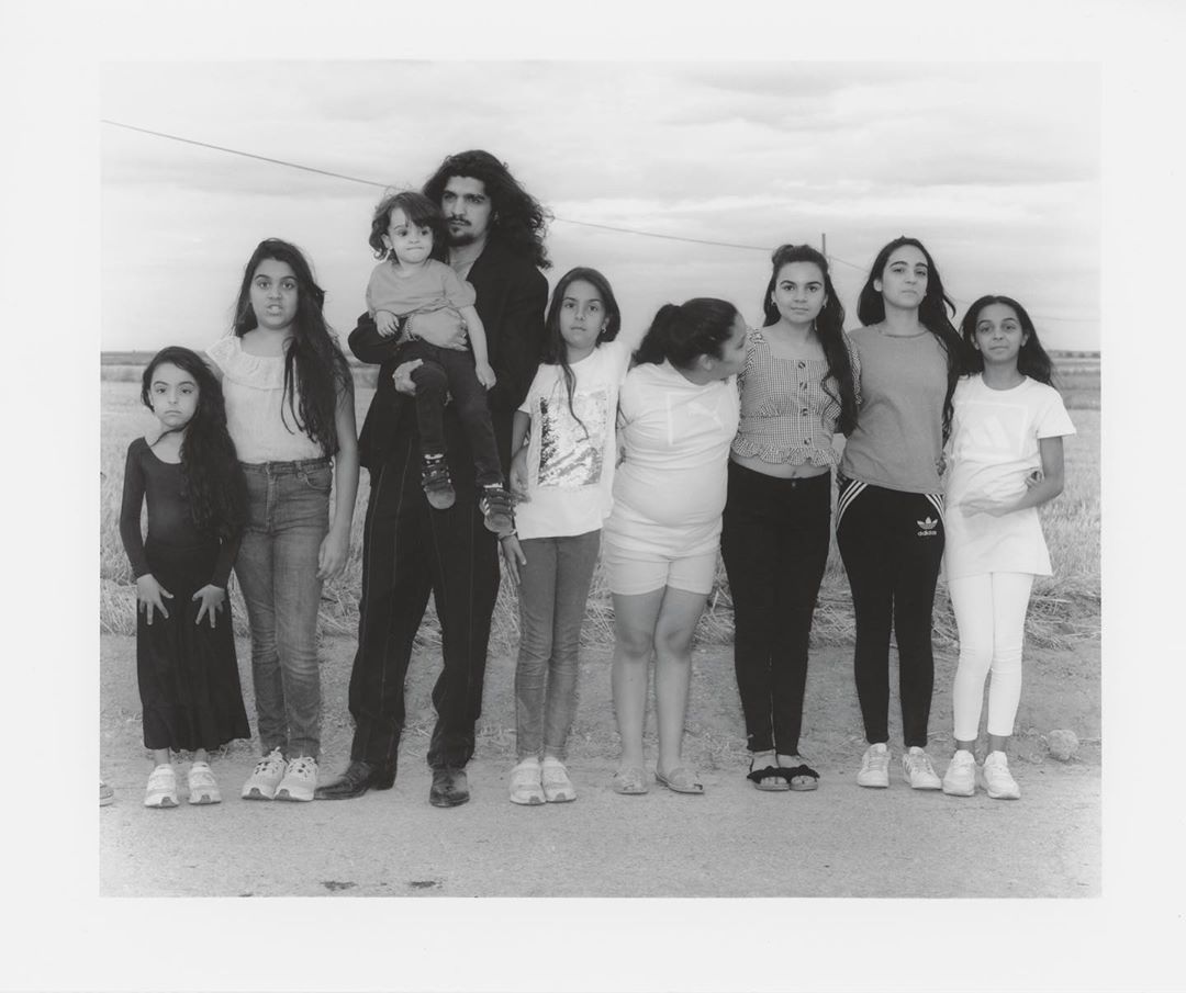 Black and white shot of a man holding a child, standing with a group of girls in an open field under a cloudy sky, in a horizontal composition.