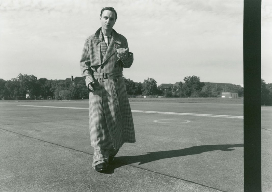 Monochrome image of a man in a patterned trench coat walking on a runway, with trees and buildings faintly visible in the background.