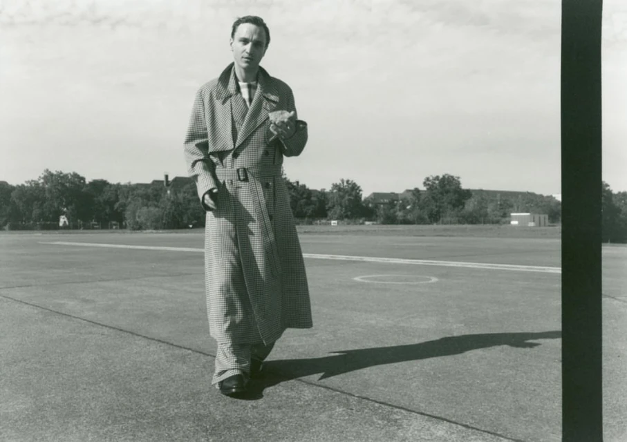 Monochrome image of a man in a patterned trench coat walking on a runway, with trees and buildings faintly visible in the background.