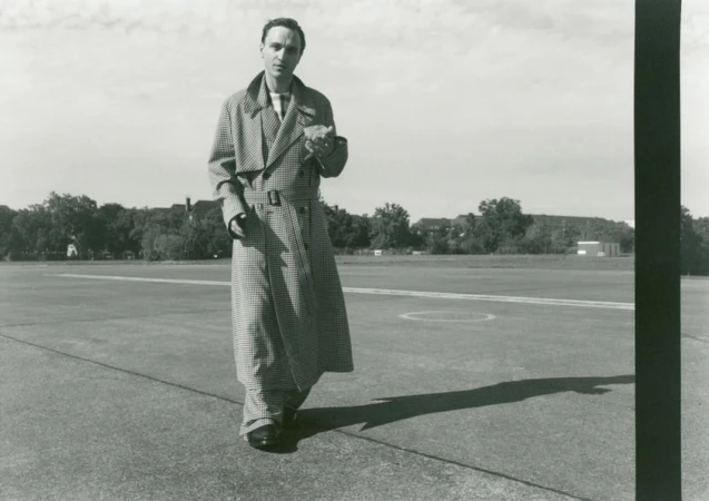 Monochrome image of a man in a patterned trench coat walking on a runway, with trees and buildings faintly visible in the background.