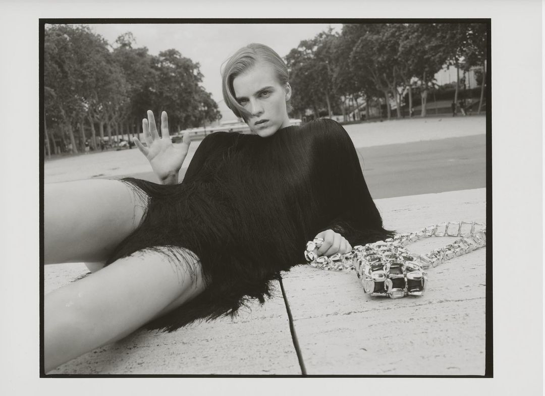 Black and white fashion shot of a female model in a fringed garment, lying on a stone surface with a silver purse, outdoors near trees.