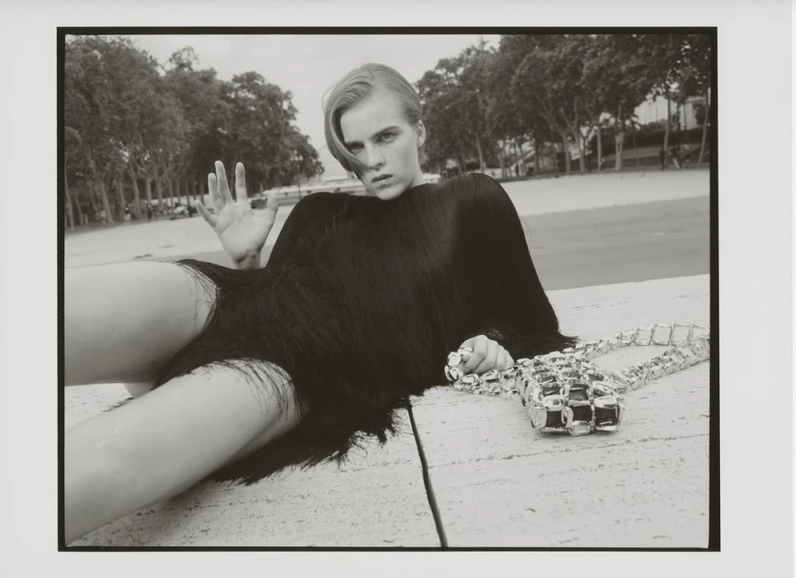Black and white fashion shot of a female model in a fringed garment, lying on a stone surface with a silver purse, outdoors near trees.