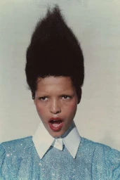 Close-up portrait of a model with towering dark hair, bleached eyebrows, wearing a sparkly blue top with a white collar against a light background.