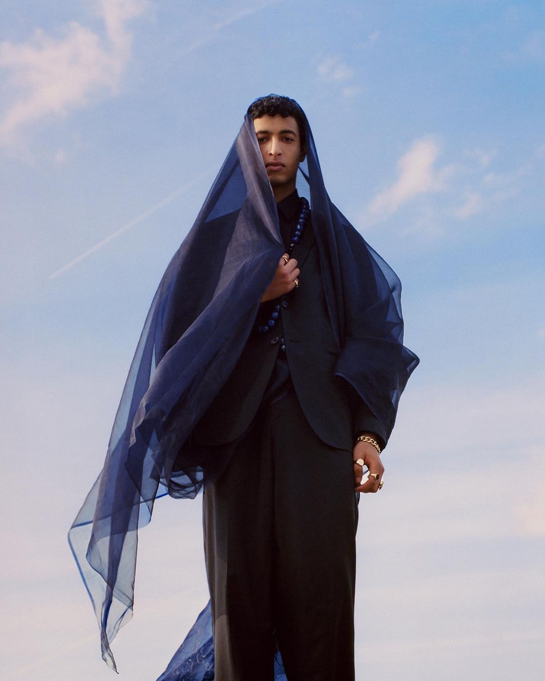 Young man in dark suit draped with a sheer blue fabric veil against a clear blue sky by Suleika Mueller