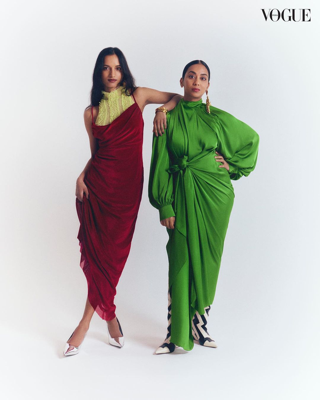 Two women pose in Vogue editorial fashion photography. One wears a red draped dress and white heels, the other wears a green dress and patterned boots.