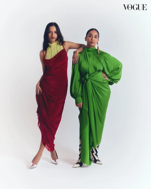 Two women pose in Vogue editorial fashion photography. One wears a red draped dress and white heels, the other wears a green dress and patterned boots.